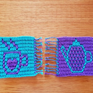 May include: Two crocheted coasters with fringe. One coaster is turquoise blue with a purple coffee cup design. The other coaster is purple with a turquoise blue teapot design.