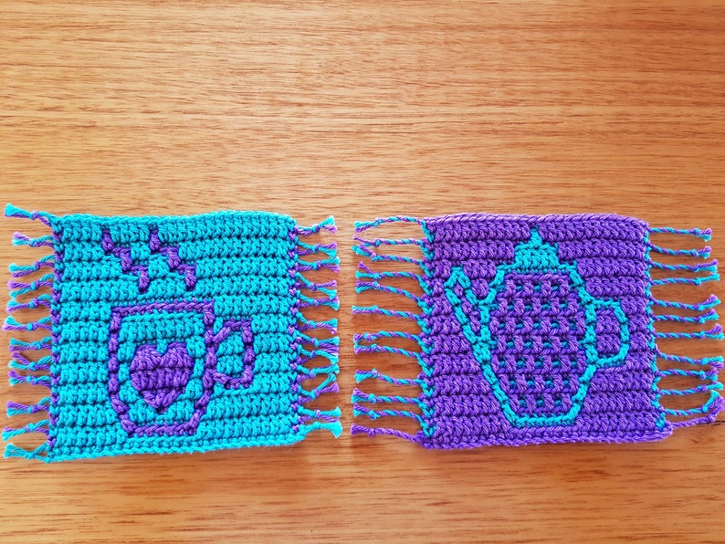 May include: Two crocheted coasters with fringe. One coaster is turquoise blue with a purple coffee cup design. The other coaster is purple with a turquoise blue teapot design.