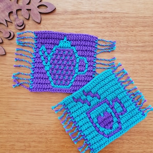 以下が含まれることがあります： フリンジが付いた2つの編み物コースター。1つのコースターは紫で、ターコイズブルーのティーポットのデザイン。もう1つのコースターはターコイズブルーで、紫の「HOT」のデザインです。