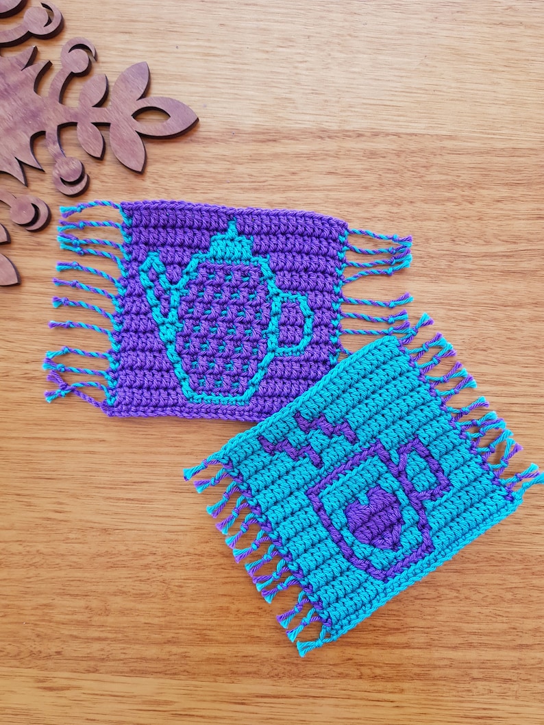 May include: Two crocheted coasters with fringe. One coaster is purple with a teal teapot design. The other coaster is teal with a purple "HOT" design.
