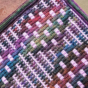 Puede incluir: Primer plano de una manta de ganchillo hecha a mano con un patrón geométrico colorido. La manta presenta una mezcla de colores que incluyen morado, verde, azul y marrón. Las puntadas de ganchillo crean una superficie texturizada.