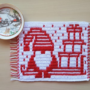 Pu&ograve; includere: Un sottopentola natalizio all'uncinetto bianco e rosso con un design di gnomo e regali. Il sottopentola ha un bordo sfrangiato ed &egrave; accanto a un piatto decorativo con una scena di Babbo Natale.