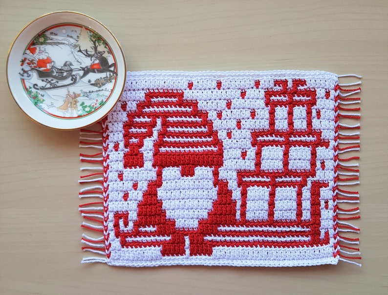 Pu&ograve; includere: Un sottopentola natalizio all'uncinetto bianco e rosso con un design di gnomo e regali. Il sottopentola ha un bordo sfrangiato ed &egrave; accanto a un piatto decorativo con una scena di Babbo Natale.