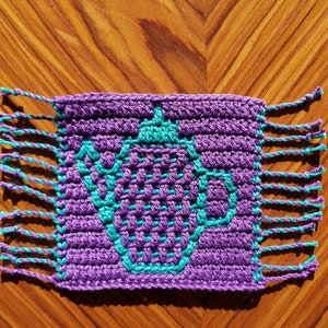 May include: Purple crocheted coaster with a teal design in the center. The coaster has fringe on all four sides.