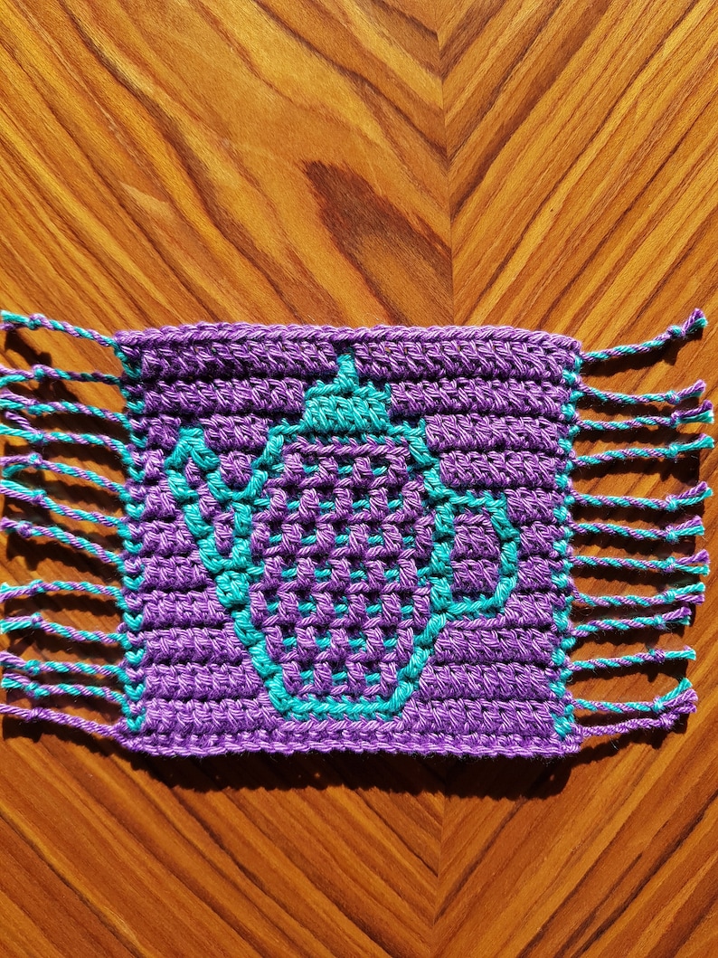 May include: Purple crocheted coaster with a teal design in the center. The coaster has fringe on all four sides.