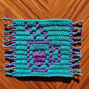 May include: A teal blue crocheted square with a purple heart design in the center. The square has fringe on all four sides.