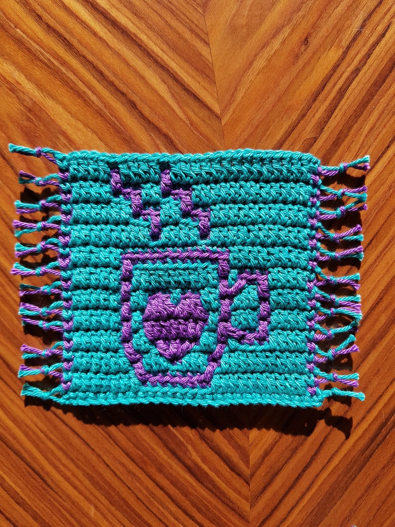 May include: A teal blue crocheted square with a purple heart design in the center. The square has fringe on all four sides.