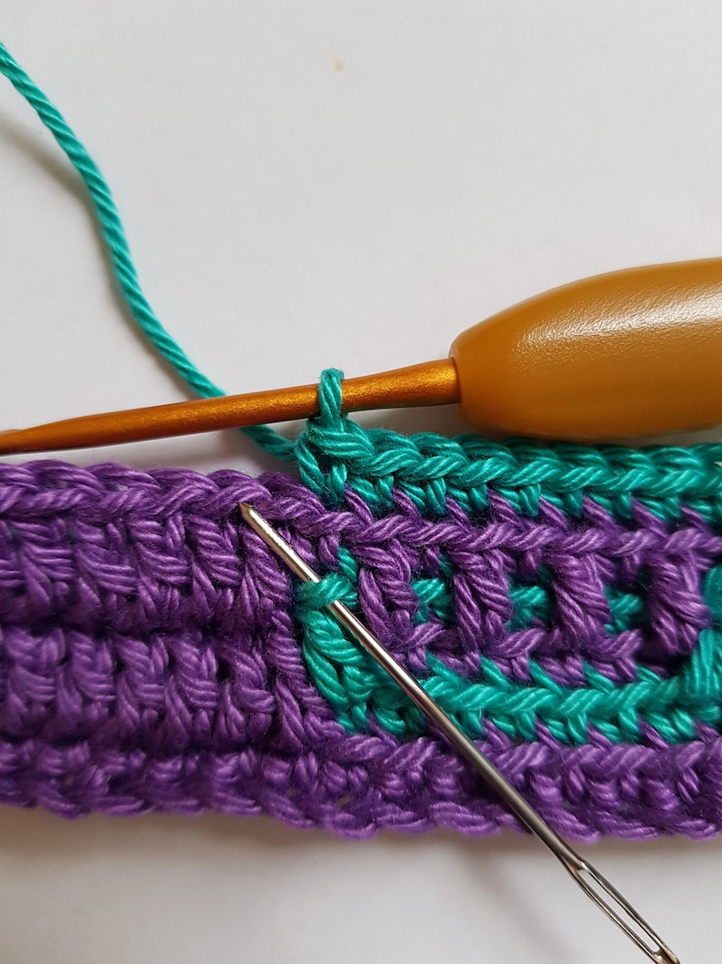 May include: A close-up of a crochet project with a purple and teal yarn. A crochet hook is being used to work a stitch in the teal yarn.