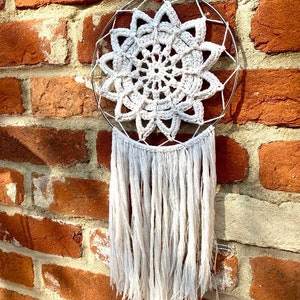 May include: A white crocheted dreamcatcher with a circular metal hoop and white fringe hanging from the bottom. The dreamcatcher is hanging on a brick wall.