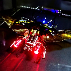 May include: A steampunk-style insect sculpture with metallic wings, gears, and red LED lights. The insect is made of metal and has a gold and black color scheme.