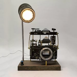 Könnte beinhalten: Eine schwarze und silberne Kameralampe mit einem Vintage-Konica-Kameragehäuse und einem schwarzen Objektiv, das an einem flexiblen Arm befestigt ist. Die Lampe steht auf einem Holzsockel und hat eine weiße Glühbirne.