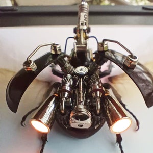 Puede incluir: Una escultura de escarabajo de estilo steampunk con intrincados engranajes de metal y un cuerpo negro. El escarabajo tiene dos grandes alas plateadas que se iluminan.