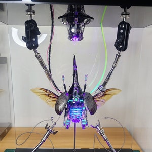 Könnte beinhalten: Eine detaillierte Skulptur eines Käfers mit mechanischen Komponenten und blau-violetten Lichtern. Der Käfer hat hellbraune Flügel und ist in einer transparenten Vitrine montiert. Die Skulptur hat silberne Metallbeine und -arme.