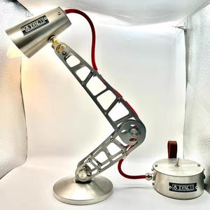 May include: A silver and red articulated desk lamp with a round base and a red cord. The lamp has a silver shade with the text "& DENJI" on it. The base also has the text "& DENJI" on it.