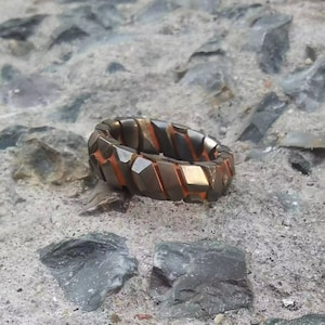 Op de afbeelding: Een zilveren ring met een getextureerd, geometrisch ontwerp. De ring heeft oranje accenten en ligt op een grijs, rotsachtig oppervlak.