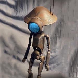 Può includere: Un robot articolato arrugginito con un grande occhio rotondo blu e una testa a cupola. Il robot è fatto di metallo e ha un aspetto invecchiato e industriale.