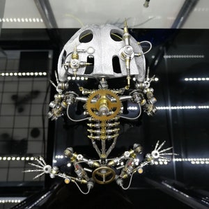 May include: A silver and gold metal sculpture of a frog with a detailed, intricate design. The frog has a helmet-like head with gears and other mechanical parts.