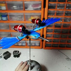 Könnte beinhalten: Eine blaue und silberne Steampunk-Vogelskulptur mit roten Motordetails. Der Vogel sitzt auf einem Metallständer und hat einen langen, spitzen Schnabel.