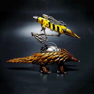 May include: A yellow and black robotic arm with a drill bit attached to the end is mounted on the back of a metal sculpture of a pangolin. The pangolin is standing on a black surface. The robotic arm has the letters "M.C.A." on the side.