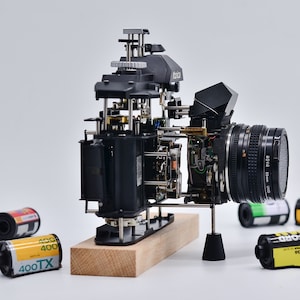 以下が含まれることがあります： 分解された黒いKonicaカメラ。レンズ付きで、木製の台座に設置。カメラの内部機構が露出しており、ギアや部品が見える。35mmフィルムのロールがカメラの周りにあり、「400TX」と表示されている。