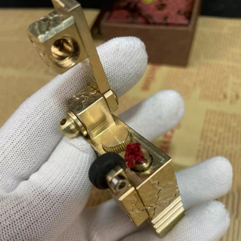 Może przedstawiać: Złoty zapalniczka z misternymi grawerunkami i czerwonym knotem. Zapalniczka ma dźwignię i koło do zapalania knota.