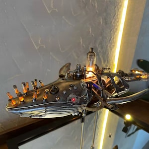 Puede incluir: Una escultura de ballena steampunk hecha de metal y engranajes. La ballena está iluminada con luces blancas cálidas y tiene un tubo de vidrio en su espalda. La escultura está montada en un soporte y parece ser una lámpara.