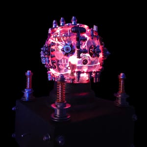 Puede incluir: Una lámpara esférica con un núcleo translúcido iluminado en rojo, adornada con numerosos pequeños componentes mecánicos y engranajes metálicos. La esfera descansa sobre una base cuadrada negra con cuatro resortes naranjas verticales en las esquinas.