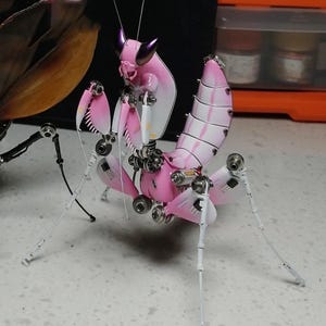 May include: A pink and white metal praying mantis sculpture with intricate details. The mantis has a segmented body and long, slender legs.