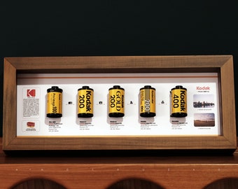 May include: A wooden frame displays a collection of six vintage Kodak film canisters. The canisters are yellow with black lettering and feature different film speeds, including 100, 200, and 400.
