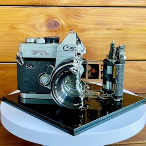 Puede incluir: Una cámara Canon FTb vintage, plateada y negra, con una lente de 50 mm, parcialmente desmontada. La cámara está sobre una superficie negra reflectante, con un fondo de madera. La lente dice "50mm 1:1.8 No.79".