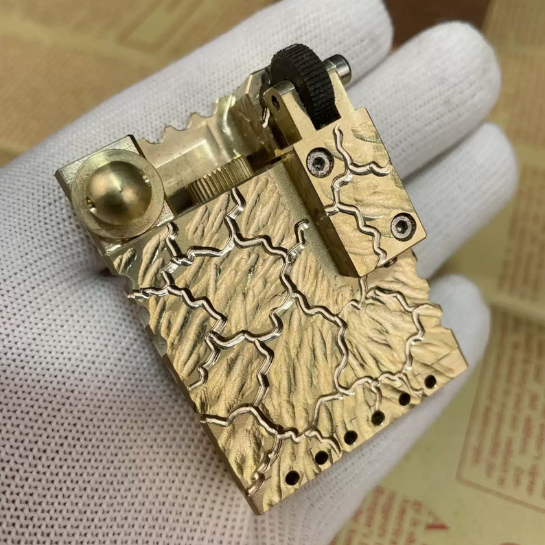 Może przedstawiać: Złoty zapalniczka z fakturowanym, wzorzystym wzorem. Zapalniczka ma mały, okrągły guzik z boku i czarne koło na g&oacute;rze.