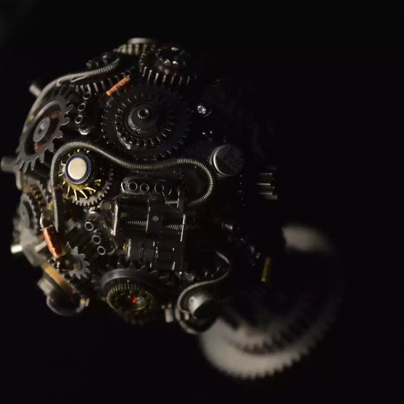Pu&ograve; includere: Primo piano di una scultura sferica in stile steampunk realizzata con ingranaggi metallici, ruote dentate e fili. La scultura &egrave; di colore grigio scuro e argento e presenta un design complesso e intricato.