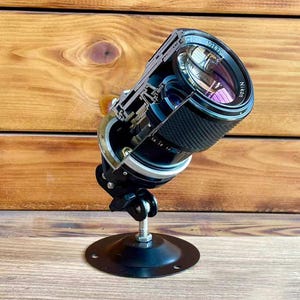 Puede incluir: Un objetivo de cámara Nikon reutilizado, cortado por la mitad para revelar sus componentes internos, montado sobre un soporte de metal negro. El objetivo tiene un agarre negro texturizado y un anillo plateado. La base es una placa de metal negra circular.