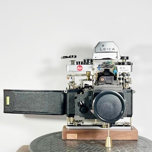 Puede incluir: Una cámara Leica R3 Electronic desmontada con un fuelle de cuero negro, una placa superior plateada con el logotipo de Leica y una base dorada. La cámara está montada sobre una base de madera con una placa de latón.