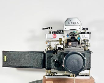 Puede incluir: Una cámara Leica R3 Electronic desmontada con un fuelle de cuero negro, una placa superior plateada con el logotipo de Leica y una base dorada. La cámara está montada sobre una base de madera con una placa de latón.