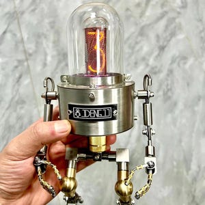 May include: A silver robot with a clear glass dome containing a red and orange Nixie tube displaying the number "3". The robot has gold-coloured arms and legs and a black label on its chest that reads "DENG LI".