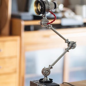 Può includere: Una lampada da scrivania unica realizzata con parti di fotocamere vintage. La lampada presenta un braccio regolabile color argento, un obiettivo della fotocamera come sorgente luminosa e un cavo rosso. La base è un classico corpo macchina nero e argento.