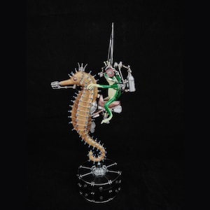 Può includere: Una statuetta di rana verde che cavalca una statuetta di cavalluccio marino marrone e blu. La rana indossa un casco e degli occhiali protettivi e tiene una lancia. Il cavalluccio marino è in piedi su una base nera.