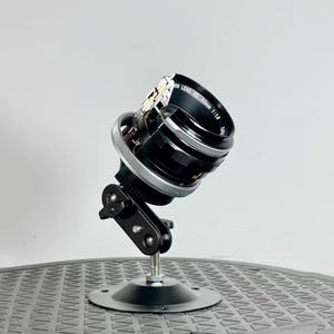 May include: A disassembled Canon FL 50mm f/1.8 lens mounted on a black tripod stand. The lens is partially disassembled, revealing the internal components.