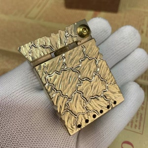 Może przedstawiać: Złoty metalowy zapalniczka z fakturowaną, wzorzystą powierzchnią. Zapalniczka ma okrągły guzik na g&oacute;rze i szereg otwor&oacute;w z boku.