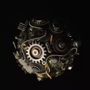 Pu&ograve; includere: Una scultura sferica in metallo con ingranaggi e ruote dentate intricati, collegati da fili e tubi metallici. La scultura &egrave; di colore grigio scuro con accenti in rame.