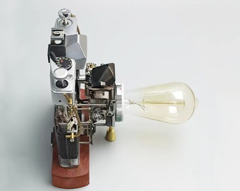 May include: A vintage camera with a light bulb attached to the side. The camera is made of metal and has a wooden base. The light bulb is clear glass with a filament visible inside.