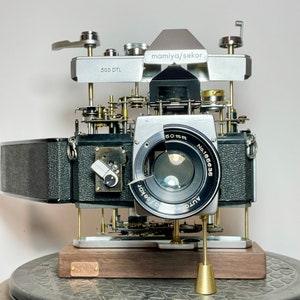 Puede incluir: Una cámara Mamiya Sekor 500 DTL plateada y negra con una lente de 60 mm. La cámara está desmontada y montada sobre una base de madera. La lente está etiquetada como "Auto Sekor 1:1.8 No.185635 60mm".