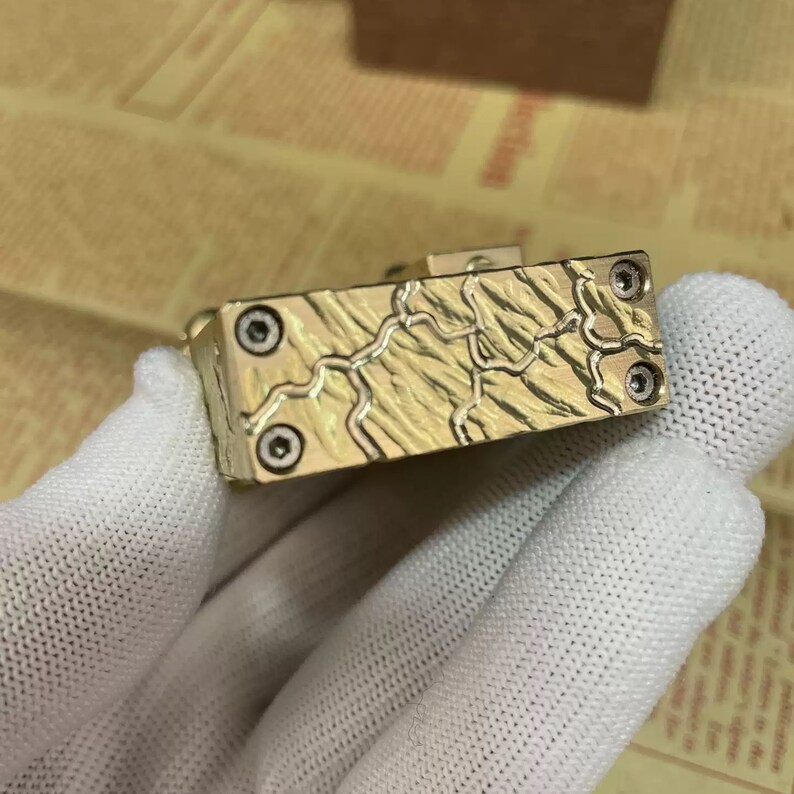 Może przedstawiać: Złoty metalowy brelok do kluczy z fakturowaną powierzchnią i czterema widocznymi śrubami. Brelok ma unikalny wz&oacute;r z liniami i krzywymi.