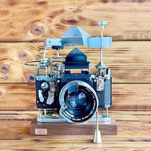 Puede incluir: Una cámara vintage negra y plateada con una lente que dice "Petri Camera" y "Auto-1:1.8". La cámara está sobre una base de madera y tiene un soporte de metal plateado con una perilla en la parte superior.