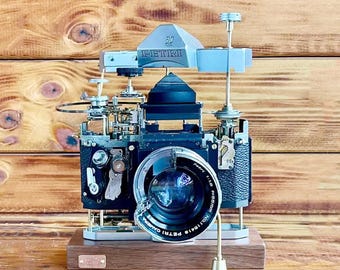 Puede incluir: Una cámara vintage negra y plateada con una lente que dice "Petri Camera" y "Auto-1:1.8". La cámara está sobre una base de madera y tiene un soporte de metal plateado con una perilla en la parte superior.
