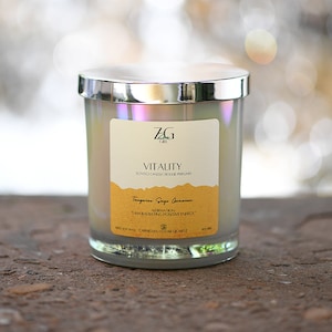 Può includere: Un barattolo di vetro per candele con un coperchio argentato. La candela è bianca e ha un'etichetta che dice "ZG Cafe Vitality Scented Candle Bougie Perfume" e "Tangerine Sage Geranium". L'etichetta include anche l'affermazione "Sto irradiando energia positiva".
