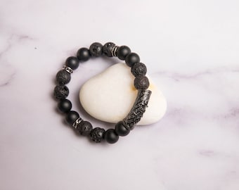 Peut inclure: Bracelet en perles d'onyx et de lave noire avec un charme en métal argenté. Le bracelet est posé sur une pierre blanche en forme de cœur.