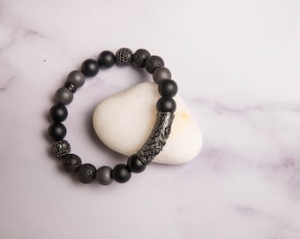 Peut inclure: Un bracelet de perles composé de perles noires mates, grises et texturées. Le bracelet comprend une perle cylindrique décorative, texturée, gris foncé. Le bracelet est présenté sur une pierre blanche et lisse sur un fond de marbre.