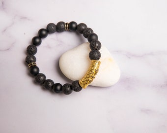 Peut inclure: Bracelet en pierres de lave noires et perles noires mates avec un charme en métal doré. Le bracelet repose sur une pierre blanche.